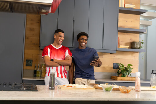 Leaning diverse male friends checking smartphone at island in modern kitchen, with pizza dough