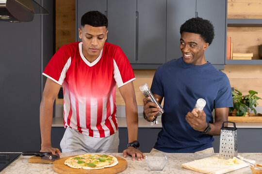 Diverse male friends seasoning pizza on marble countertop in kitchen, with salt and pepper grinders