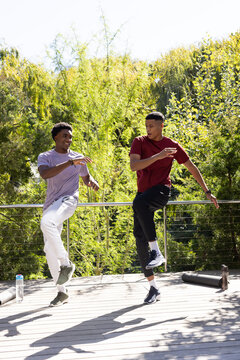 Dancing diverse male friends executing aerobic moves on wooden deck outdoors, with water bottles