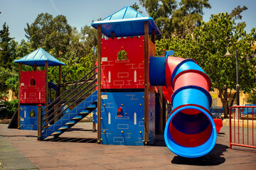 Bright and colorful castle-themed playground with a spiral tube slide, stairs, and towers. Outdoor recreational area for children surrounded by trees and benches, on a sunny day.