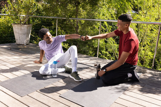 Fistbumping Diverse male friends stretching on yoga mats on wooden deck terrace, with water bottles