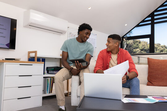 Diverse male coworkers discussing printouts on sofa in office, with tablet and laptop