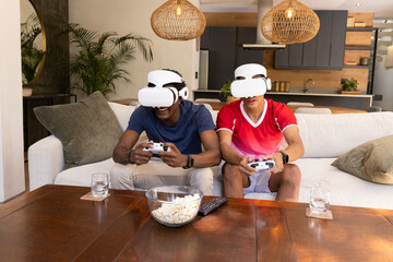 Diverse male friends playing virtual reality game on sofa in living room, with headsets and popcorn