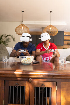 Playing diverse male friends using VR headsets on sofa in living room, with popcorn, controllers