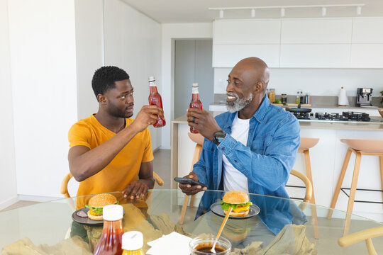 African American father and son celebrating lunch in kitchen, holding red bottles and hamburgers