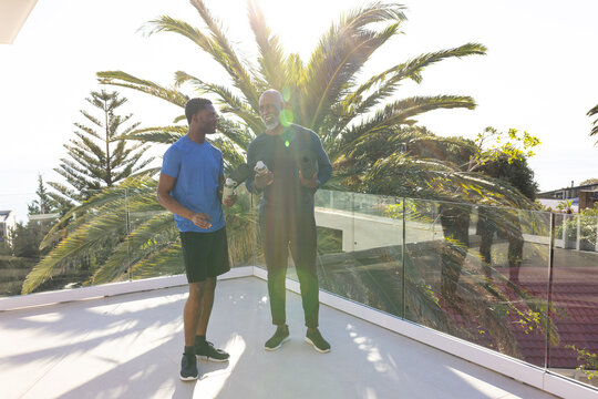 Standing African American father and son holding water bottles on rooftop terrace, with foam roller