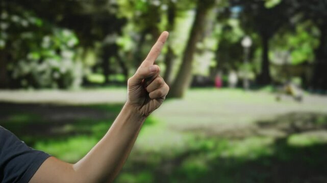 Man's hand pointing fingers upward against blurred green park background in a sunny outdoor setting.