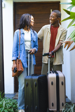 Smiling African American couple standing on front porch, holding suitcases and smartphones