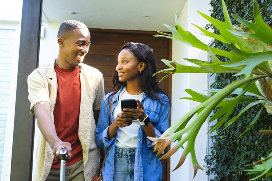 Smiling African American couple posing at front door, holding suitcase smartphone and backpack