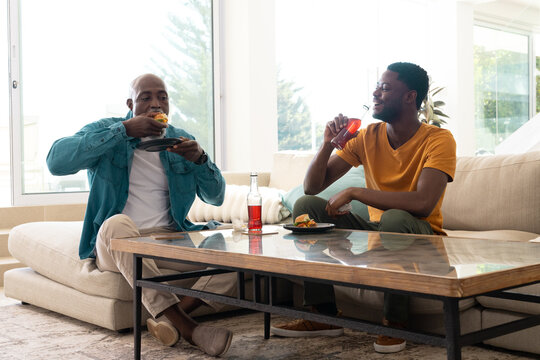 African American father and son eating burgers and sipping soda in living room, with smartphone