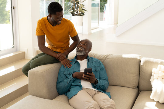 Reclining African American father and son holding smartphone on beige sectional sofa in living room - Powered by Adobe