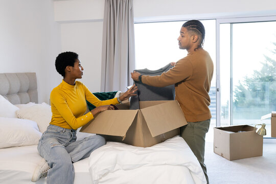 Diverse couple unpacking clothes from cardboard box on bed in bedroom, with pillows