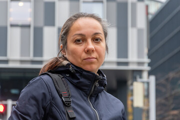 A serious 40-45-year-old woman with gathered dark hair looks directly at the camera against the background of a modern building with large windows.
