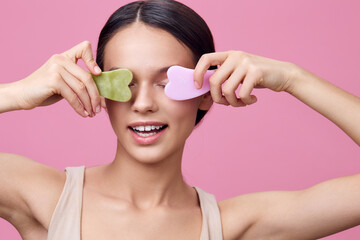Young woman using jade rollers on her face, smiling joyfully against a pink background, promoting...