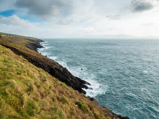 A beautiful ocean view with a rocky cliff in the background. The water is choppy and the sky is cloudy. Dingle peninsula, county Kerry, Ireland. Popular tourist area with stunning nature scenery.