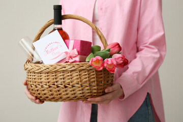 Young woman holding basket with wine bottle, tulips and gift box for Mother's Day on white...