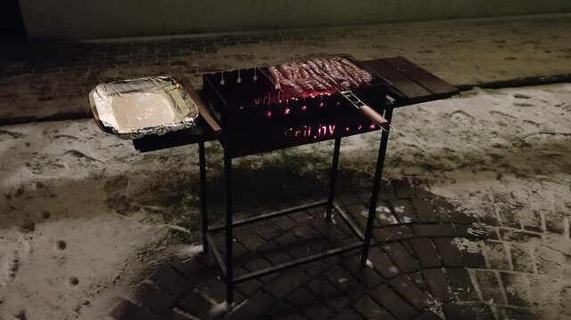 In the yard, a metal brazier stands on a tiled area next to a lawn and the wall of the house. Coals lie under a wooden-handled grate filled with mullet fish, the fire burning faintly. Snow has fallen
