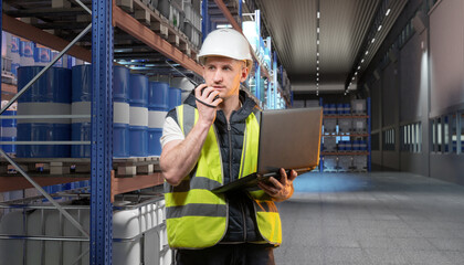 Warehouse supervisor with laptop and walkie-talkie checks inventory of chemical barrels on...