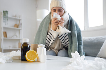 Different flu medications with lemon on table against sick woman sneezing at home, closeup