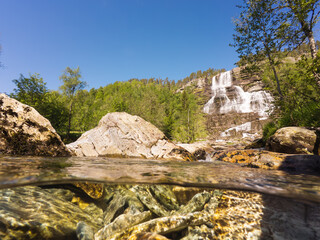 Famous Tvindefossen waterfall in Norway: summer sunny landscape with a unique over-under view of clear water, rocks, and green forest.