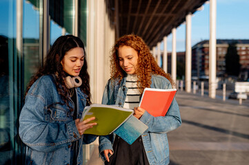 Two young latin women sitting together on campus, reviewing class notes and discussing study materials before attending their next class