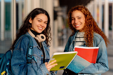 Two happy latin female students carrying notebooks and backpack returning to university campus