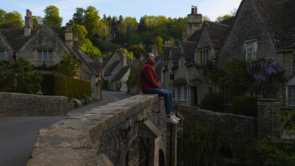 Man sitting on a bridge of a charming fairytale village in the Cotswolds, England