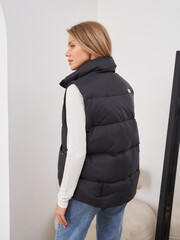 Woman with long hair in black puffer vest and white top, denim jeans, stands beside a wall and...