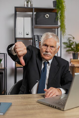 Dislike. Upset elderly businessman working on laptop at home office thumbs down sign gesture,...
