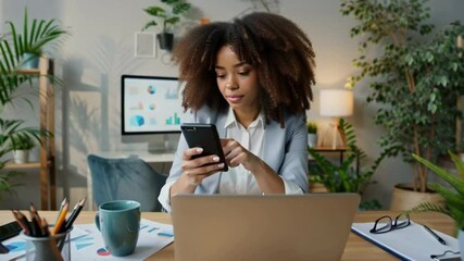 Confident young adult african american businesswoman using smartphone and laptop while working remotely in cozy home office filled with lush plants and natural light - Powered by Adobe