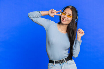Happy woman in headphones listening music and dancing disco fooling around having fun expressive gesticulating hands relaxing on party. Brunette girl in sunglasses isolated on studio blue background