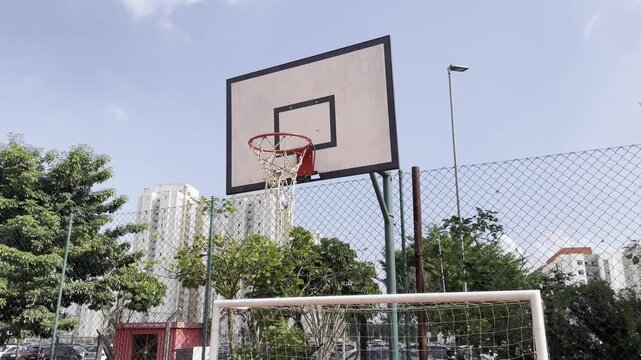 Basketball hoop above a five-a-side soccer goal. Old and dirty outdoor basketball hoop. White clouds, blue sky, green vegetation, old red court, chain link fence. 4K HDR 30fps.