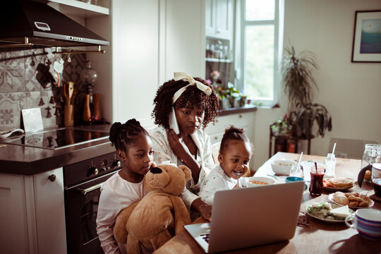 Single mother having breakfast with her two daughters while talking on the phone and using a laptop at home