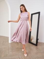 Woman in a flowing pink sleeveless dress with white polka dots poses gracefully indoors. She stands...