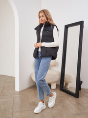 Young woman in casual outfit poses indoors, wearing a black puffer vest, cream sweater, jeans, and...