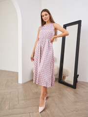 Elegant woman in a sleeveless, pink polka dot dress poses confidently indoors, standing next to a tall mirror on wooden flooring. She wears light-colored heels and has long hair © Dmitry Tsvetkov