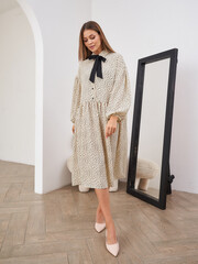 Woman with long hair wearing a beige and black polka dot dress with a black bow tie, standing...