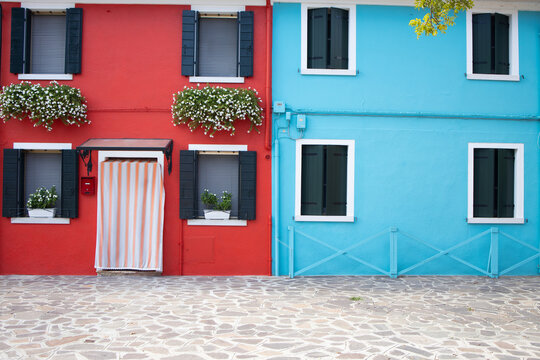 Vivid red and turquoise blue house facades side by side in Burano, Italy. The red house features black shutters, white flower boxes, and a striped curtain on the door. The bright colors and neat desig