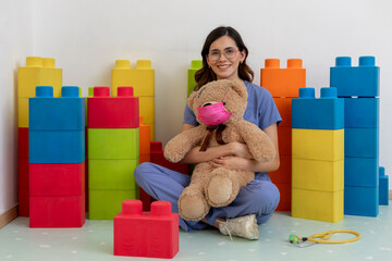 Pediatrician wearing scrubs holding teddy bear with mask in doctor's office