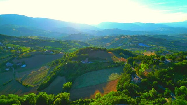 Relaxing aerial push-in shot from San Ginesio, the balmy hillscapes in its outskirts bathed in suffused afternoon sunlight, gently strewn thickets, fields, punctuated buildings, Sibillini mountains