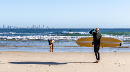 A surfer with board looking towards the water from Coolangatta Beach with the high-rise city skyscape of the Gold Coast on the horizon.