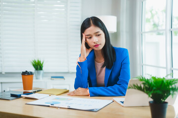 Asian businesswoman in a blue blazer sits with eyes closed and hand on temple, showing signs of...