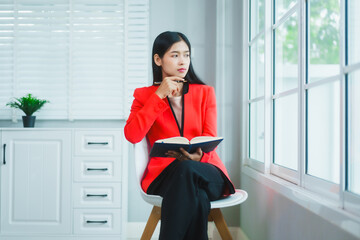 Confident Asian businesswoman smiles while talking on the phone beside a window in a modern office,...