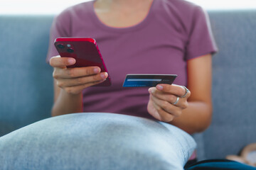 Happy Asian woman sits comfortably on a sofa,smiling while holding multiple credit cards,representing convenient online shopping,smart financial planning,modern digital consumption in a relaxed home