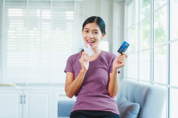Happy Asian woman sits comfortably on a sofa,smiling while holding multiple credit...