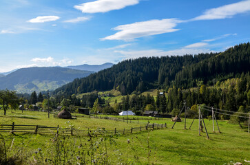 Peaceful rural setting with distant mountains, dense forests, and green open pastures. Scenery includes wooden fences and small cottages, creating a tranquil and picturesque atmosphere. Carpathians