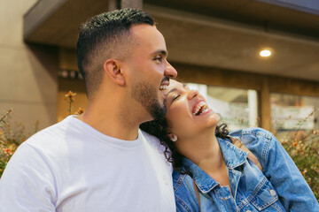 Young latin woman rests her head on her boyfriend shoulder, both laugh on their date night outdoors