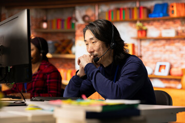 Call center worker dealing with upset customers, speaking with them online using headset at office desk. Asian man using PC to do customer support job in workplace, handling clients complaints