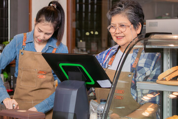 Young attractive barista working together with senior elderly retirement woman while using POS...