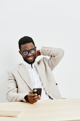 Smiling happy young man with afro hairstyle wearing glasses and beige blazer sitting at desk using smartphone, isolated on plain white background. Casual, cheerful, adult, elearning, technology